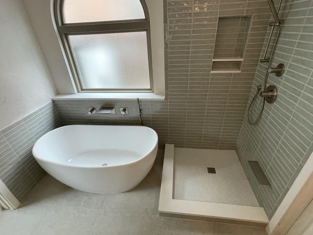 Gray-tiled bathroom with a freestanding tub and walk-in shower beside an arched window